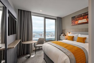 A hotel room with a large bed, a desk, a chair, a TV mounted on the wall, and a city view through the window.