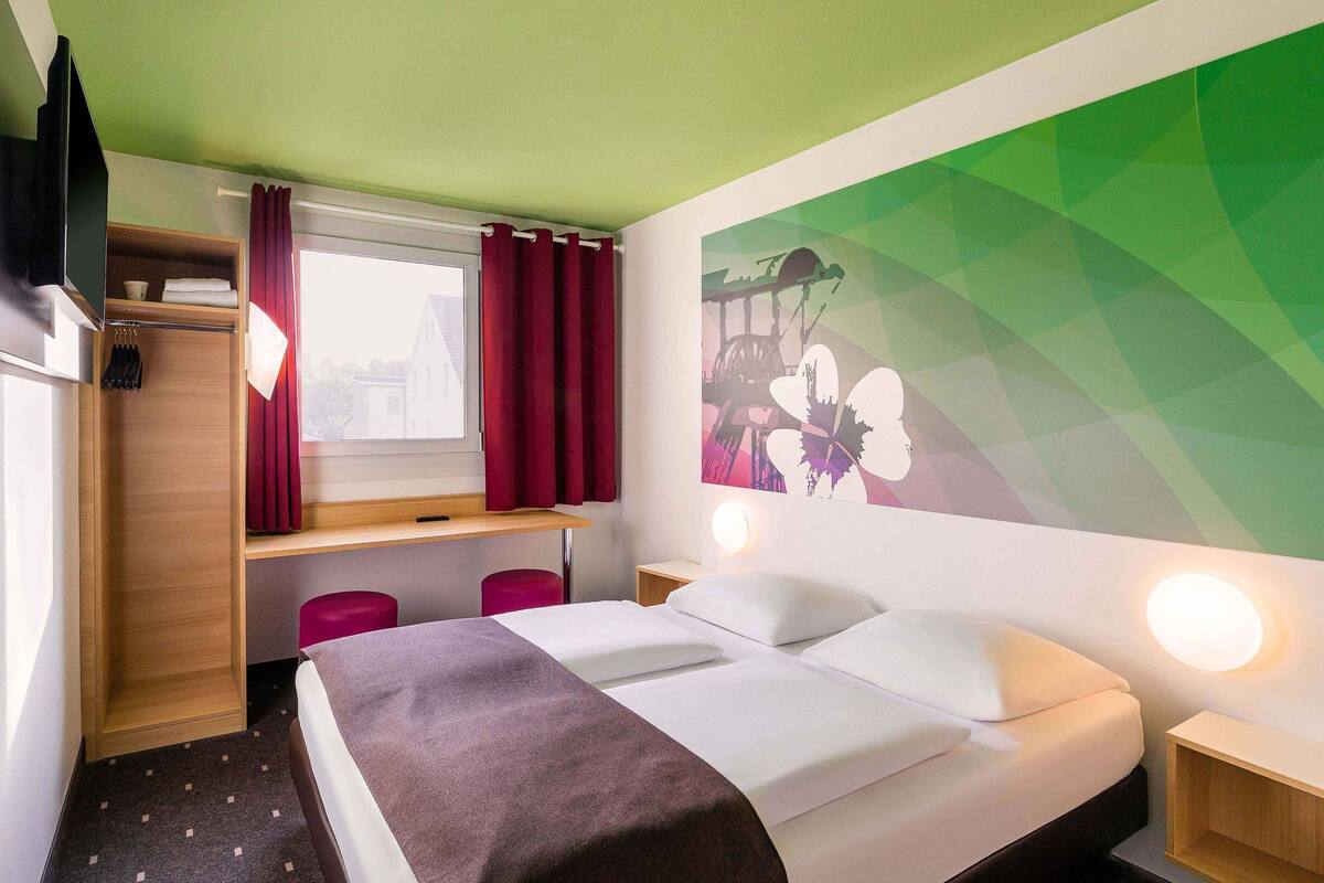 Habitación de hotel con cama, escritorio, silla, armario y ventana con cortinas.