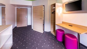 Une chambre d’hôtel avec un plafond vert, une porte en bois, une armoire, un téléviseur fixé au mur, un petit bureau en bois et un tabouret violet.