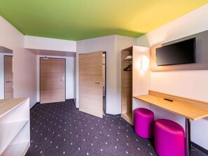 Une chambre d’hôtel avec un plafond vert, une porte en bois, une armoire, un téléviseur fixé au mur, un petit bureau en bois et un tabouret violet.
