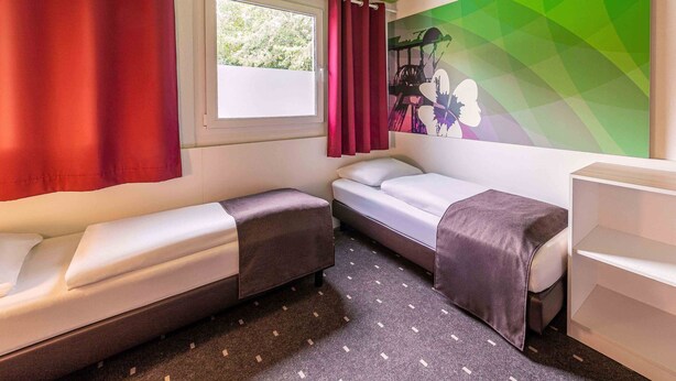 Une petite pièce bien éclairée, avec deux lits simples, une fenêtre avec des rideaux, un mur orné d’une fresque géométrique et un bureau blanc.