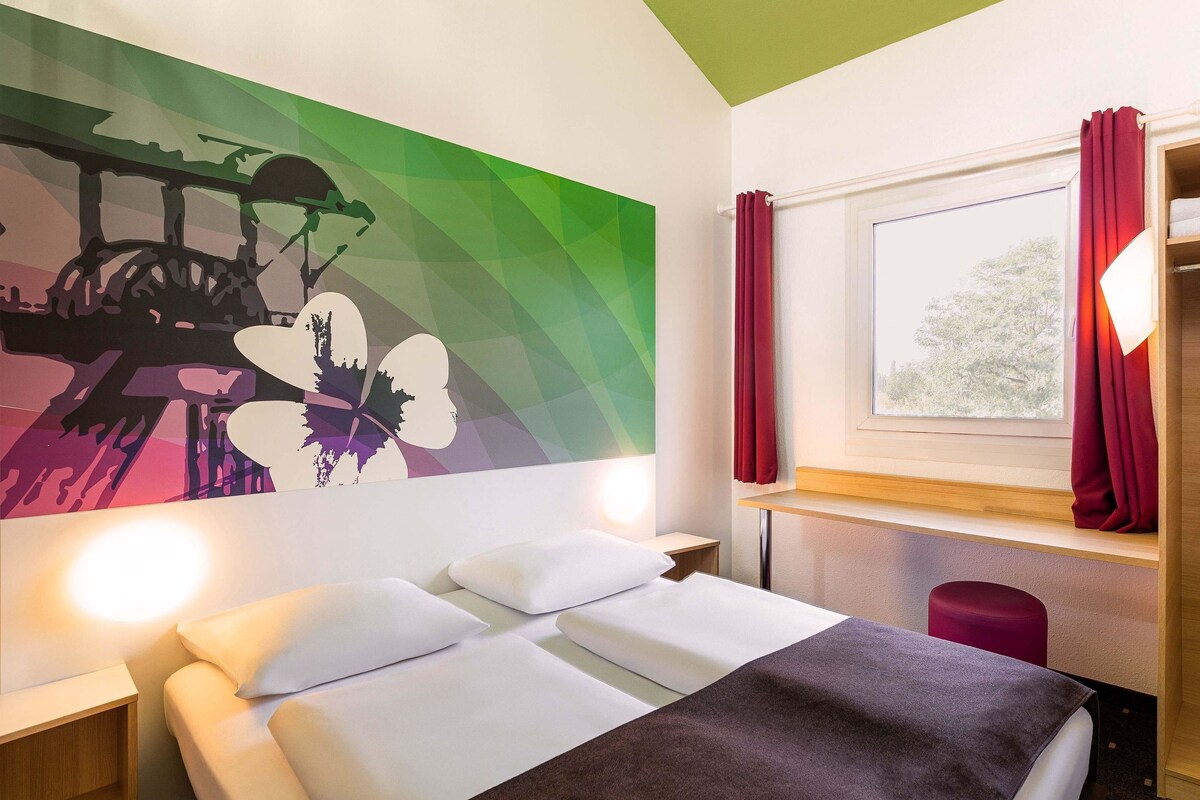 Ein Hotelzimmer mit Bett, Nachttisch, Fenster mit Vorhängen und Wand mit farbenfrohem Wandbild.