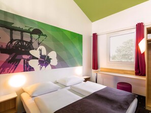 Una habitación de hotel con cama, mesita de noche, ventana con cortinas y una pared con un mural colorido.