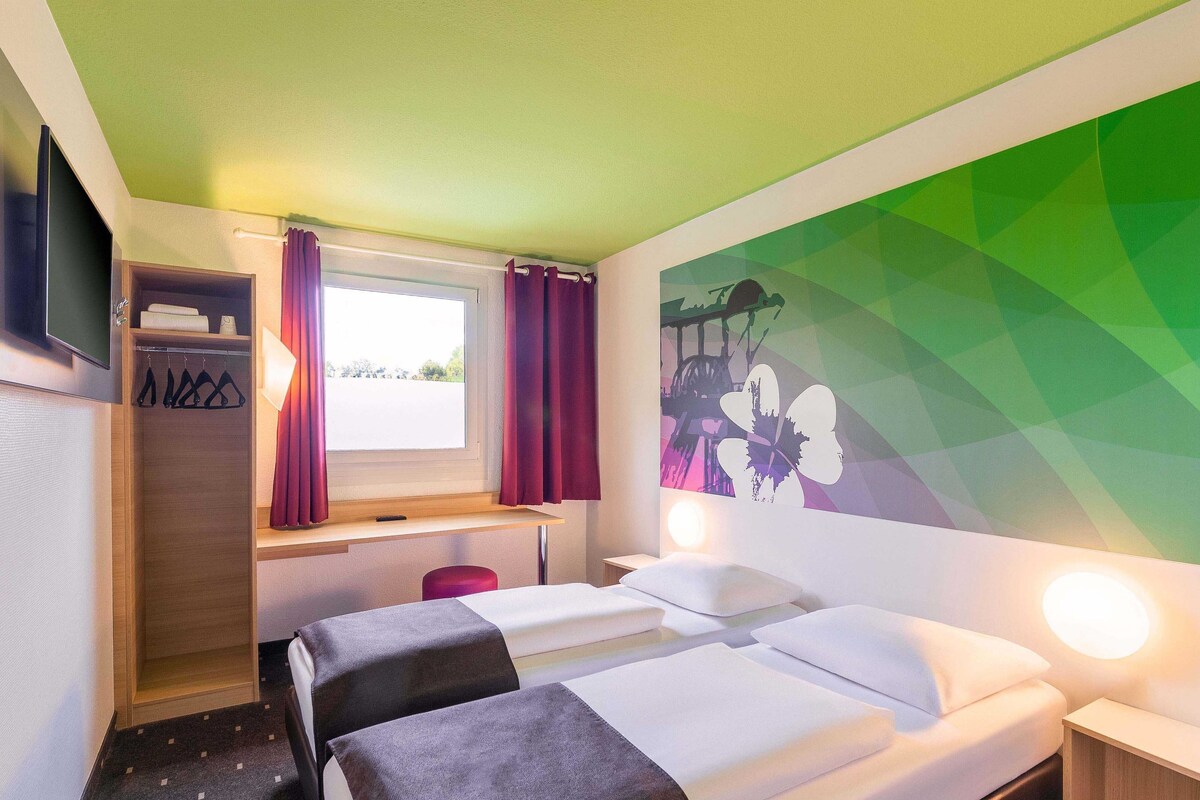 Ein Hotelzimmer mit einem Bett, einem Schreibtisch, einem Kleiderschrank und einer Wand mit einem geometrischen Blumenbild.