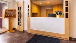 Une salle d’attente avec un bureau en bois, un ordinateur et une plante. On y trouve un comptoir d’enregistrement et un panneau d’affichage où sont accrochées des brochures.