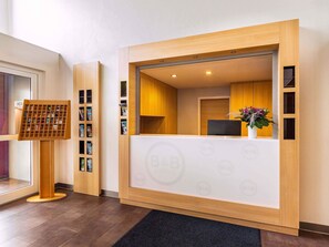 Ein Empfangsbereich mit einem Holztisch, einem Computer und einer Pflanze. Es gibt eine Rezeption und ein Schwarzes Brett mit Broschüren.
