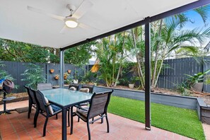 Enjoy a peaceful outdoor space with a glass-top table and comfortable seating for four, surrounded by lush greenery and a ceiling fan.