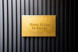 A gold plaque with "Guest house by hosoda" engraved on it, mounted on a wall with vertical slats.