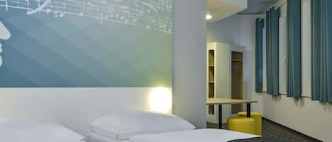 Habitación de hotel con dos camas individuales, un televisor montado en la pared, una mesita con un taburete amarillo y una ventana con cortinas.