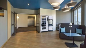 A modern lobby with a wooden floor, a vending machine, a sofa, and a staircase leading to an elevator. - B&B HOTEL Halle (Saale) (Halle (Saale))