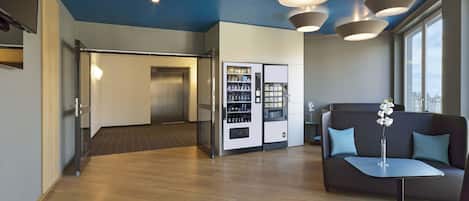 Um hall moderno com piso de madeira, uma máquina de vendas, um sofá e uma escada que dá acesso a um elevador.