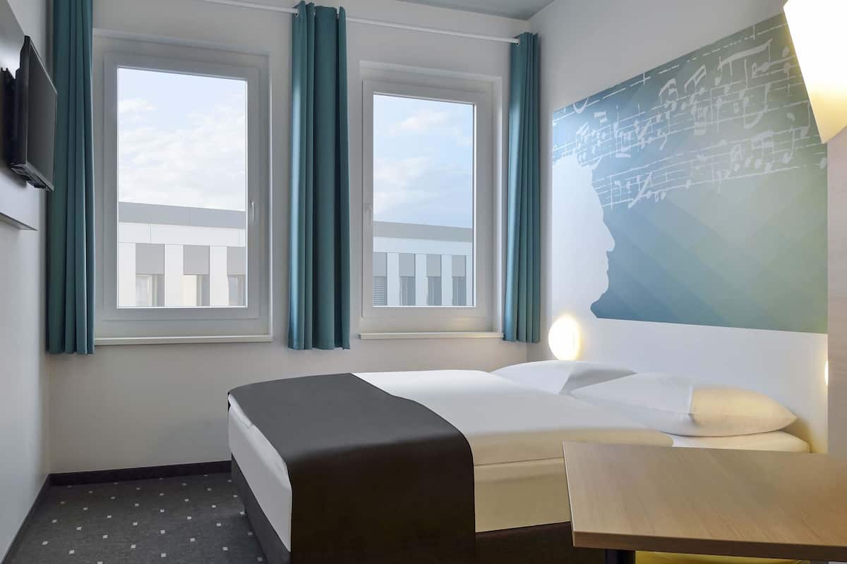 Habitació d'hotel amb un llit, una taula de treball i dues finestres amb cortines.