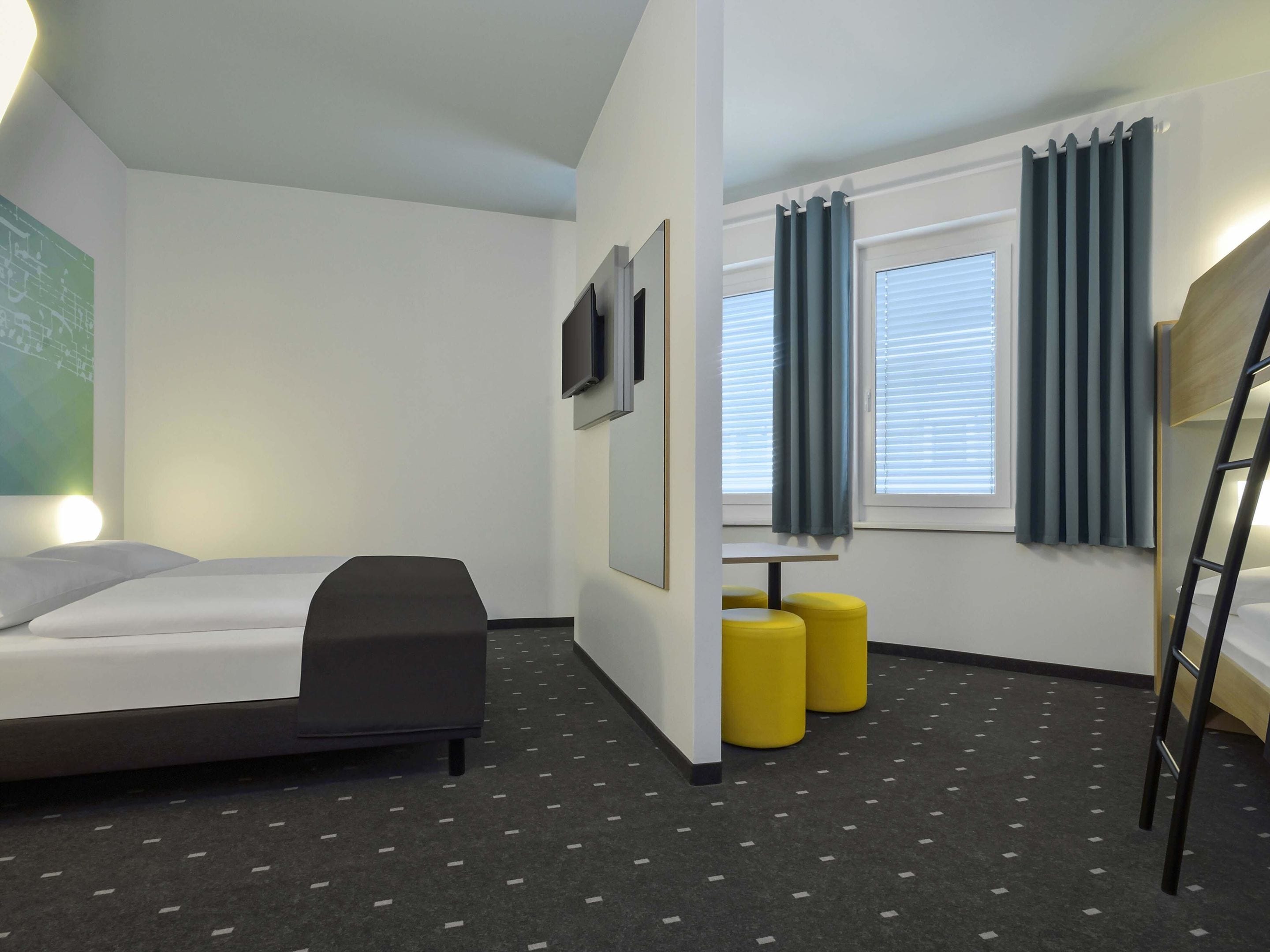 Una habitación de hotel moderna con una cama litera, un escritorio, dos otomanas amarillas y un televisor montado en la pared.