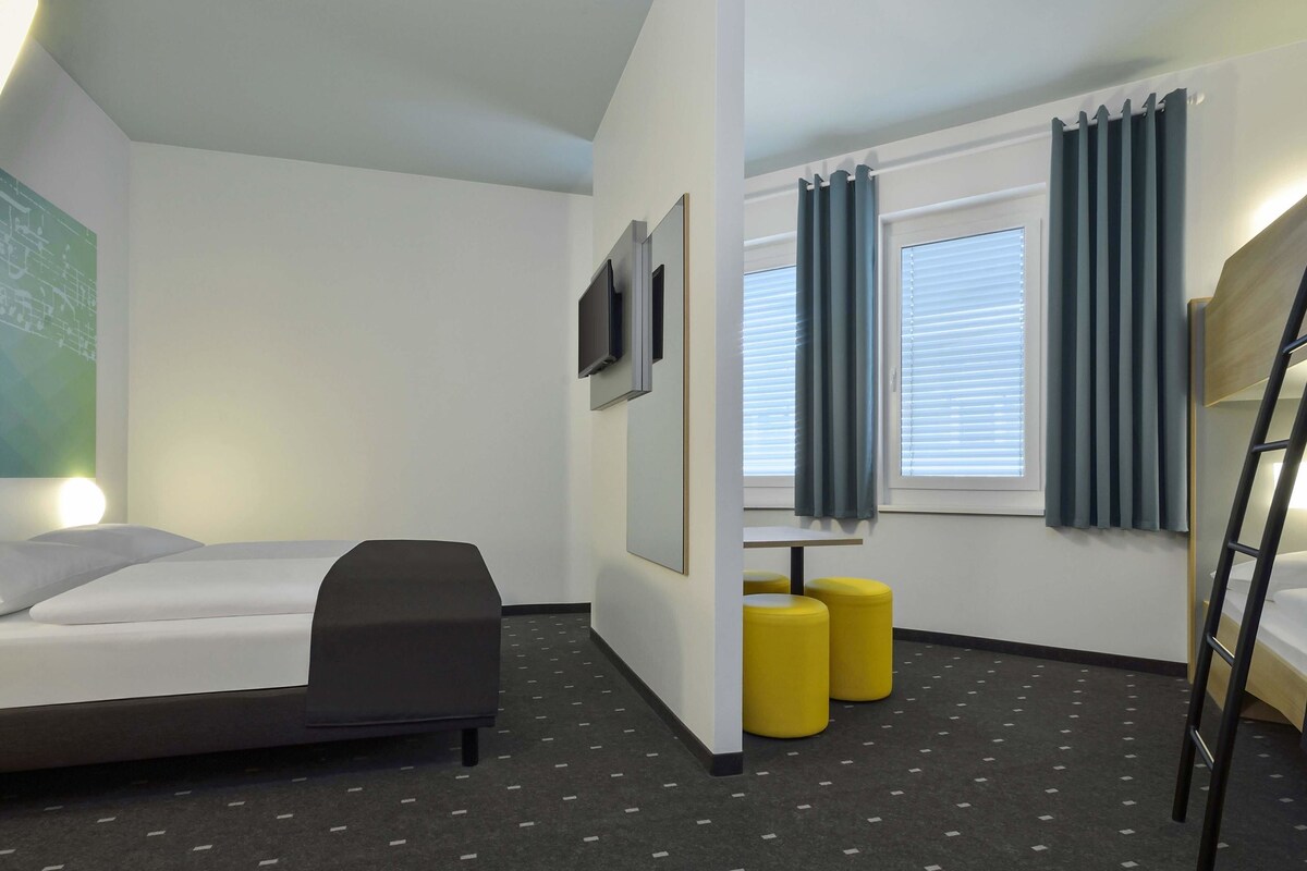 Una habitación de hotel moderna con una cama litera, un escritorio, dos otomanas amarillas y un televisor montado en la pared.