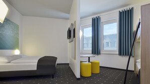 A hotel room with a bed, a desk, two yellow ottomans, a TV, and a window with curtains. - B&B HOTEL Halle (Saale) (Halle (Saale))