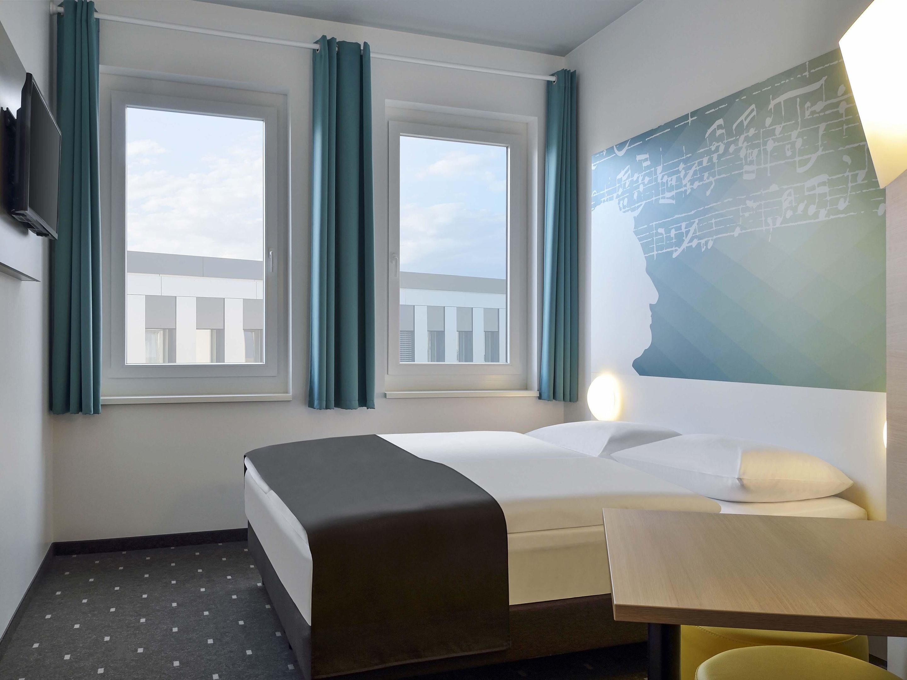 A hotel room with a bed, a desk, and two windows with curtains.