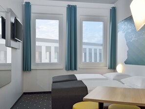 Habitación de hotel con cama, escritorio, silla, televisor y ventanas con cortinas.