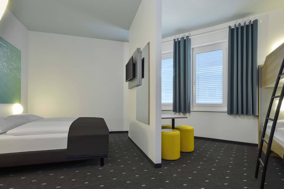 Habitació d'hotel moderna amb un llit superposat, un escriptori, dos otomans grocs i un televisor fixat a la paret.