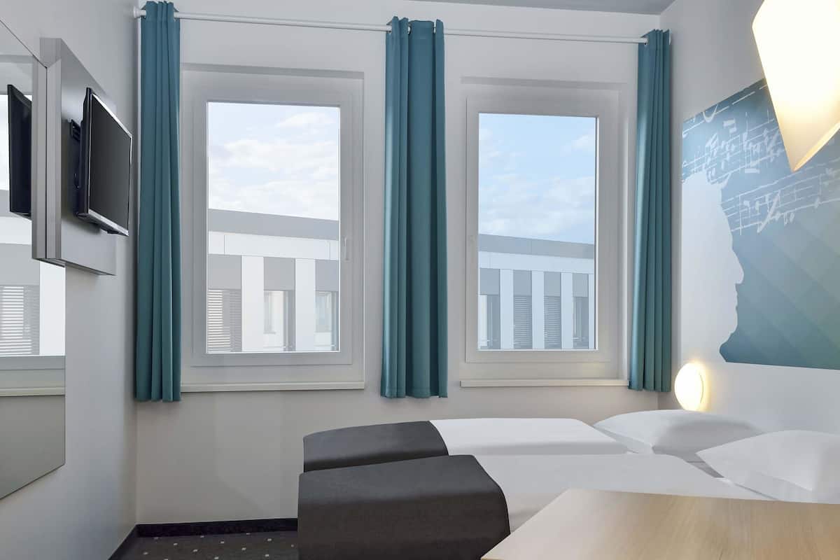Habitació d'hotel amb un llit, escriptori, cadira, televisió i finestres amb cortines.
