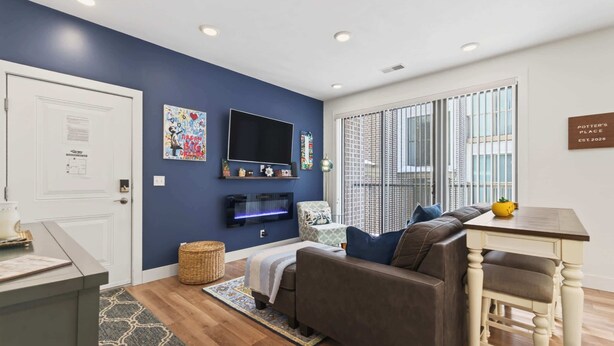 A living room with a sofa, a TV mounted on the wall, a window with blinds, and a sign on the wall.