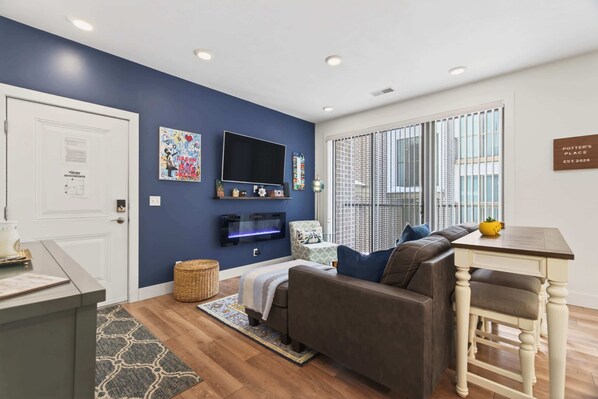 A living room with a blue accent wall, a flat-screen TV, a sofa, a dining table, and a window with blinds.