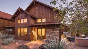 A two-story wooden house with a stone foundation, a patio with outdoor furniture, and a desert landscape. - Coyote Run - Cozy Rustic Retreat in Moab- Private Hot Tub-Fireplace (moab)