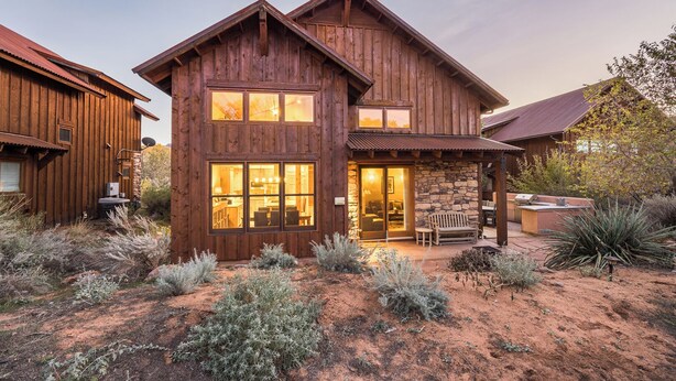 A rustic wooden house with a stone foundation, large windows, and a patio area with outdoor seating. - Coyote Run - Cozy Rustic Retreat in Moab- Private Hot Tub-Fireplace (moab)