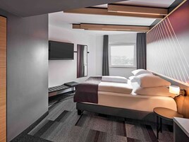 A modern hotel room with a large bed, a desk, a flat-screen TV, and a window with curtains.
