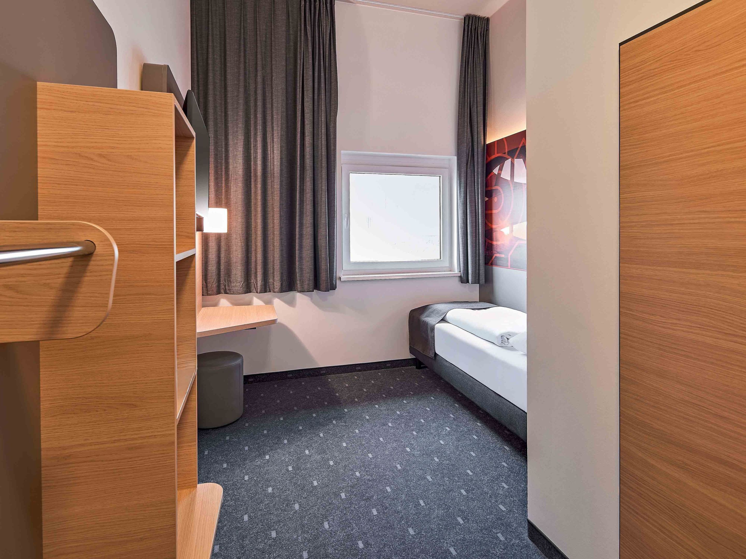 A compact hotel room with a bed, a small window with curtains, a wooden wardrobe, and a trash bin.