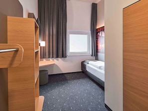 A compact hotel room with a bed, a small window with curtains, a wooden wardrobe, and a trash bin. - B&B Hotel Hamm (Hamm)