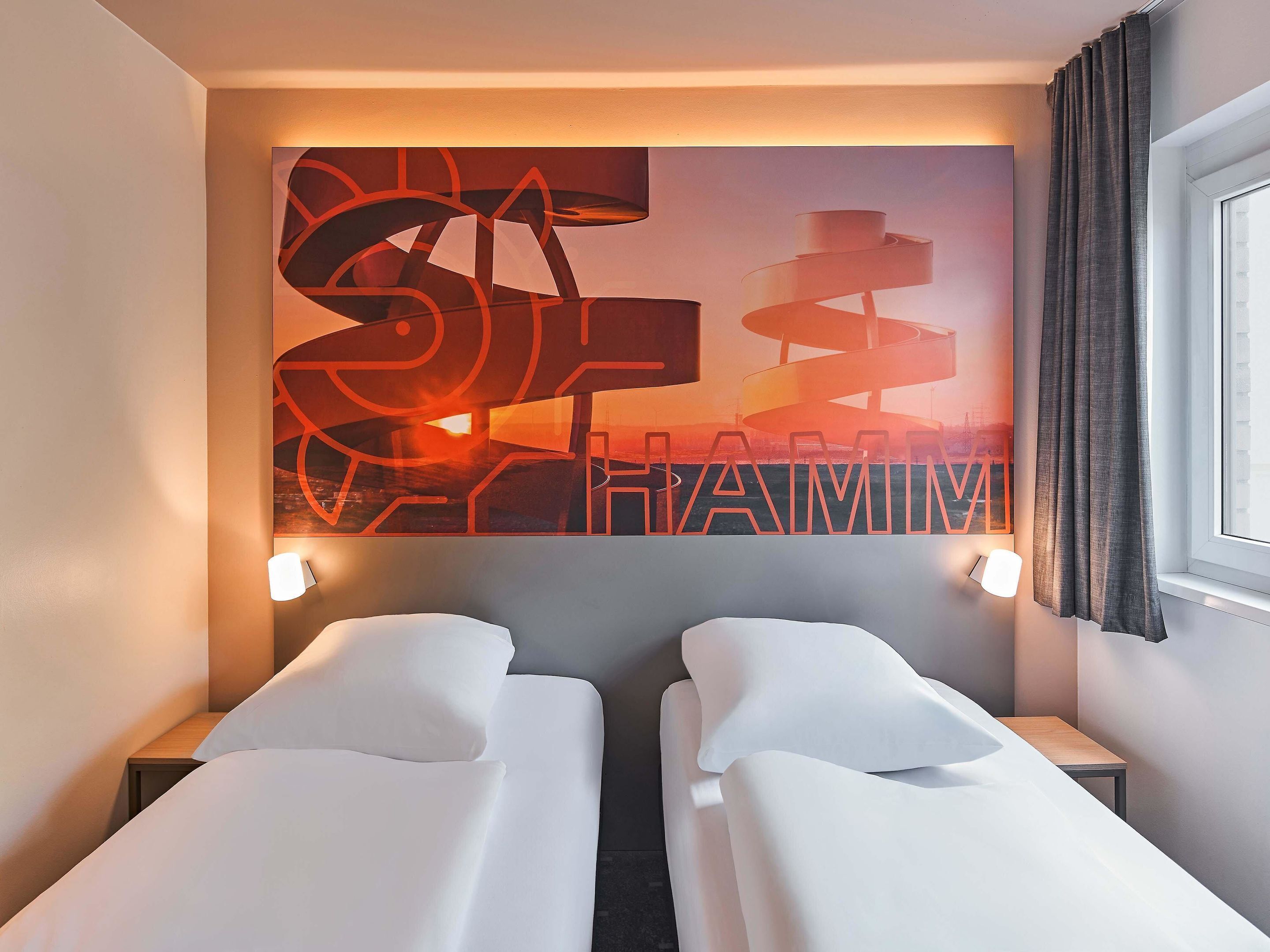 Una camera d'albergo con due letti, un quadro appeso al muro e una finestra con tende.