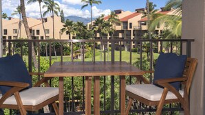 Een balkon met twee houten stoelen en een tafel, met uitzicht op een aangelegde tuin met palmstruiken en een blik op hotelgebouwen.