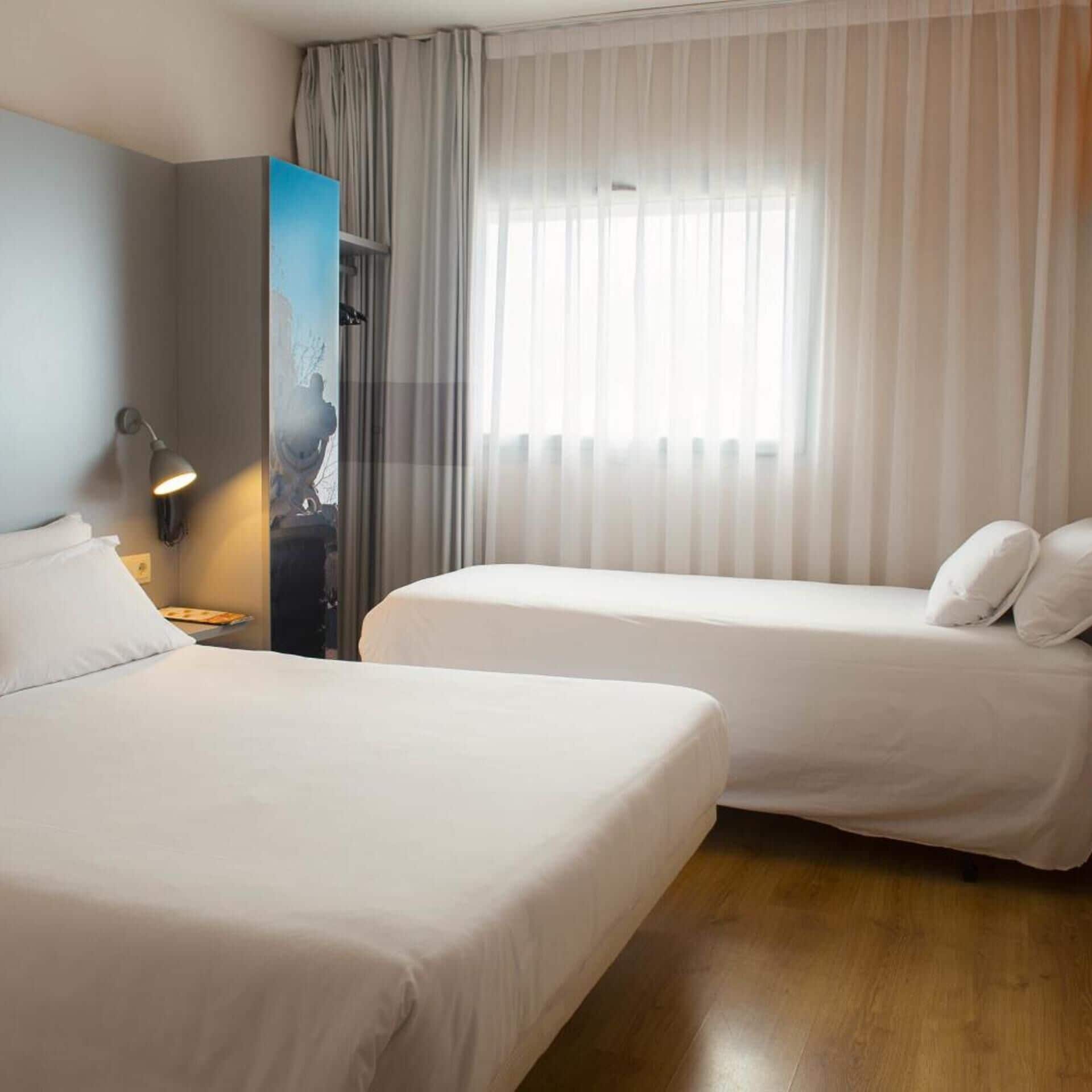 A hotel room with two beds, a nightstand with a lamp, a wardrobe, and a window with sheer curtains.