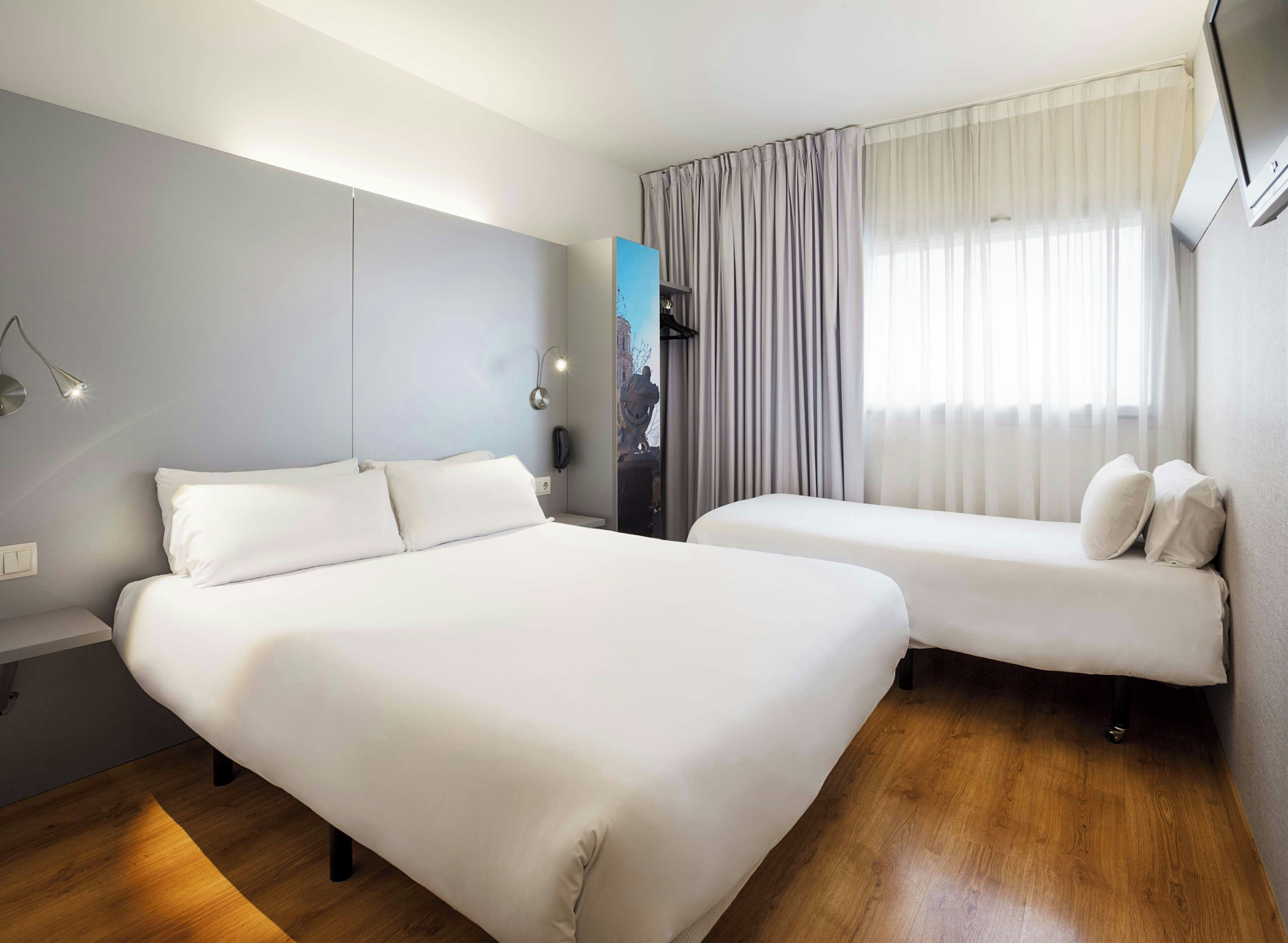 Une chambre d’hôtel avec un grand lit, un lit plus petit, une table de chevet, une lampe et une fenêtre avec des rideaux.