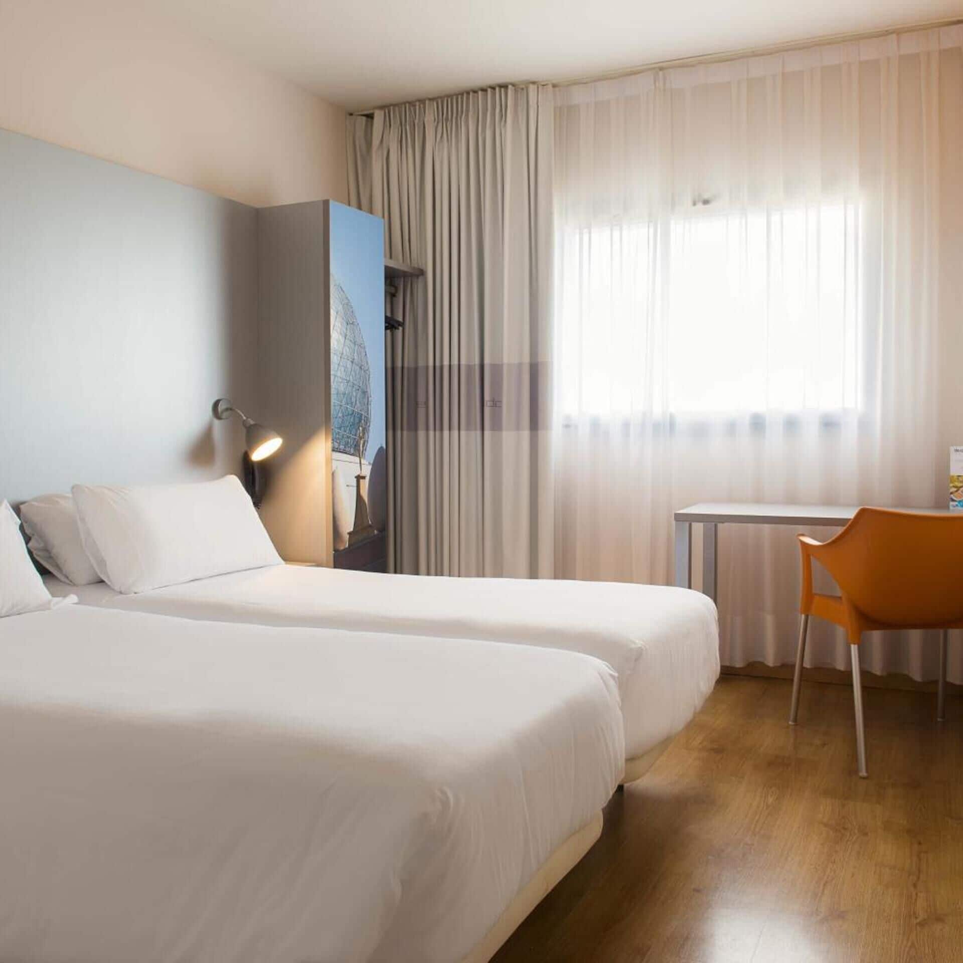 Ein Hotelzimmer mit zwei Betten, einem Schreibtisch, einem Stuhl, einer Lampe und einem großen Fenster mit Vorhängen.