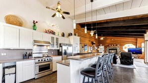 A modern kitchen with white cabinetry, a central island with bar stools, a stone fireplace, and a large TV in the living area. - Tahoe's Most Sought After Lakeview -Cliff Hanger Home less than 1 Mile to Heavenly, Sleeps 10 (South Lake Tahoe)