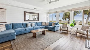 Uma sala de estar com um sofá seccional azul, uma mesa de centro de madeira e vista para o oceano através de grandes janelas.