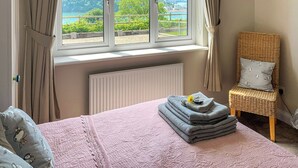 Une chambre avec une grande fenêtre, un lit recouvert d’une couette rose, une chaise avec un coussin et un radiateur.
