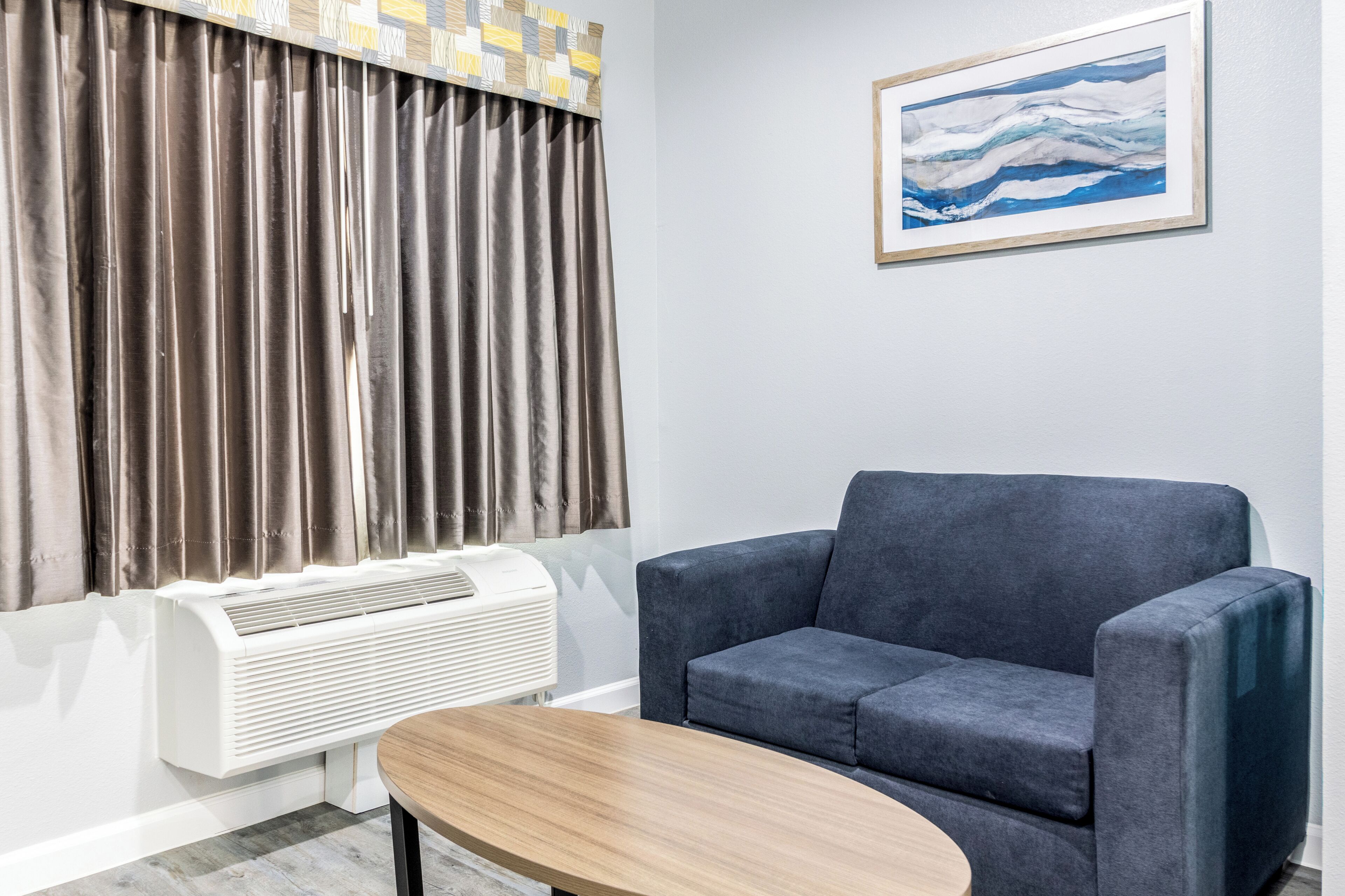 A room with a gray sofa, a wooden coffee table, a white air conditioning unit, and a painting on the wall.
