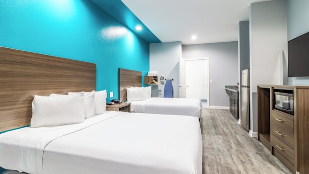 Une chambre d’hôtel avec deux lits, une tête de lit en bois, une kitchenette et une salle de bain visible au fond.