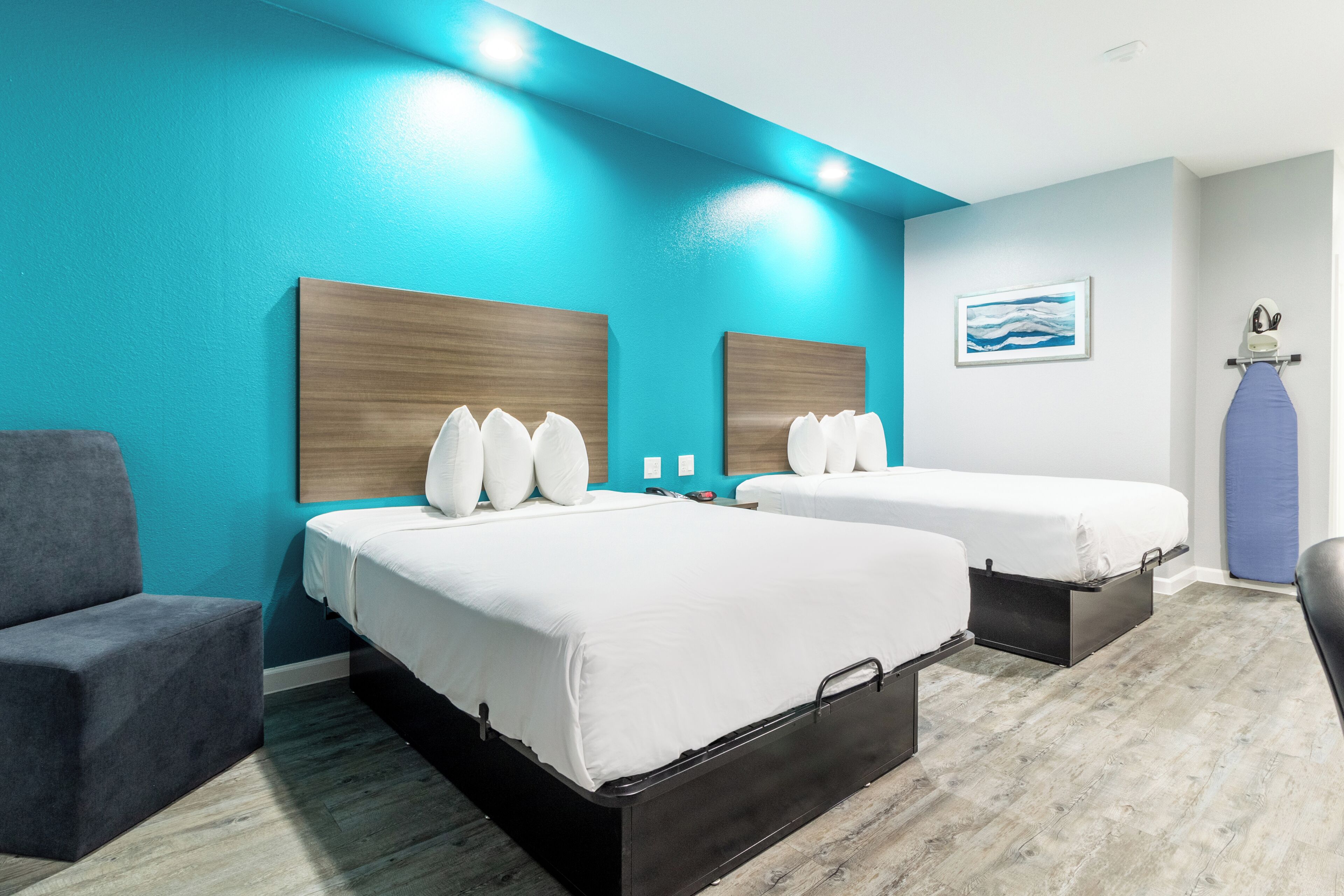 A hotel room with two beds, a gray armchair, a blue accent wall, and a painting on the wall.