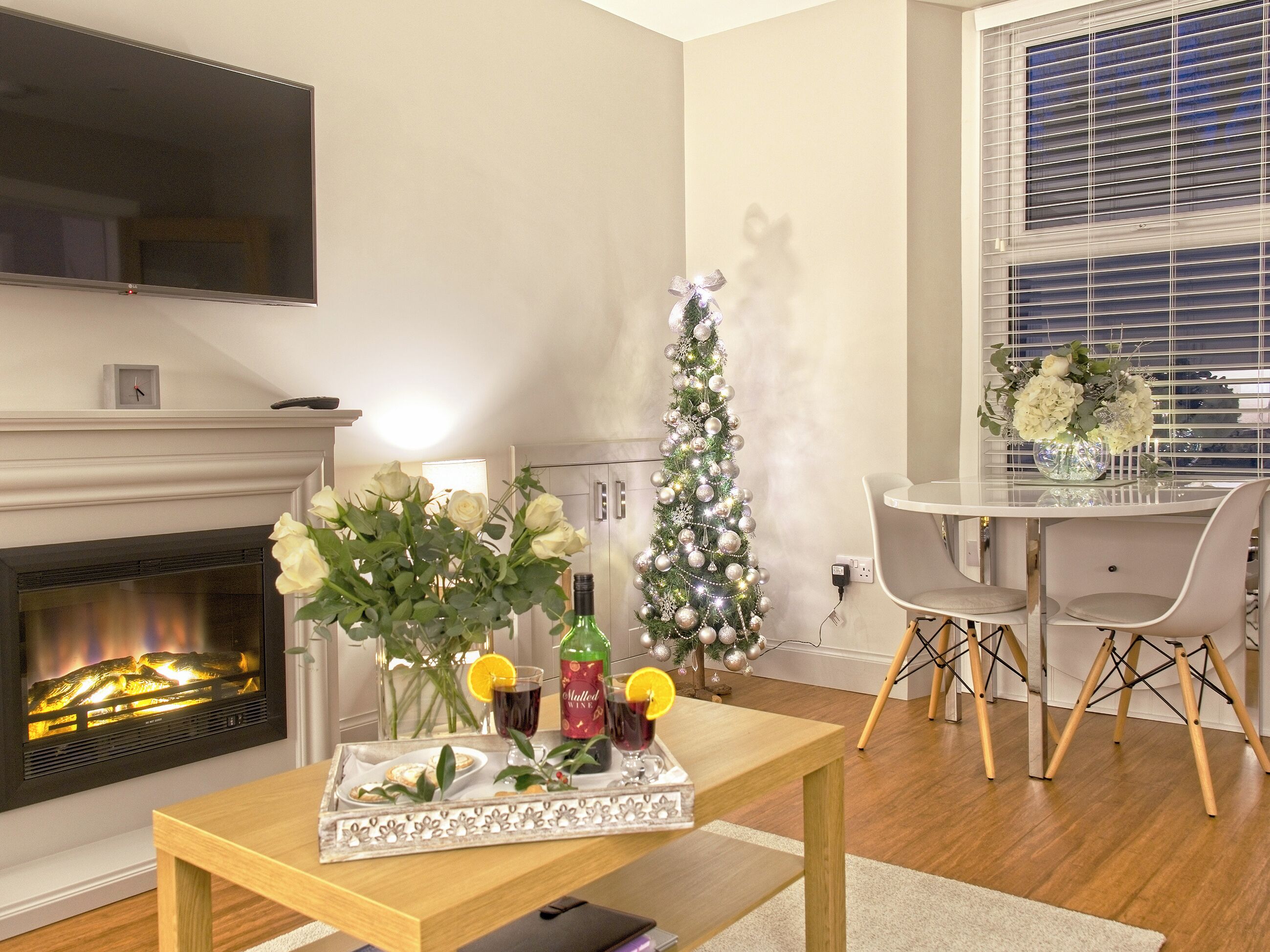 Un salon avec une cheminée, un sapin de Noël, une table à manger avec des chaises et un téléviseur fixé au mur.