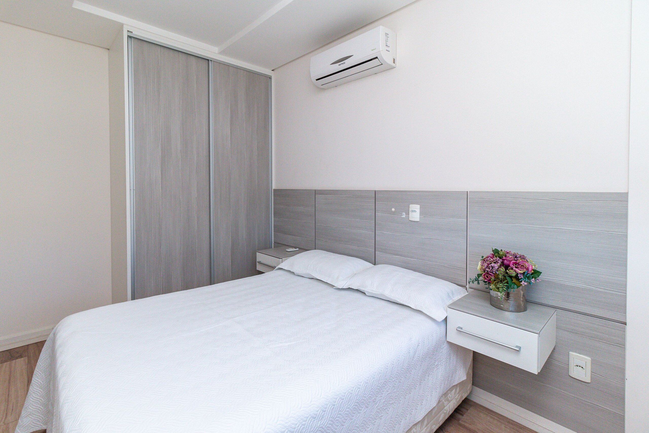 Un dormitorio con cama, mesitas de noche, aire acondicionado y una estantería de pared con un jarrón de flores.