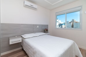 Une chambre à coucher comprenant un lit, une table de chevet, une commode, un climatiseur et une fenêtre donnant sur des immeubles.