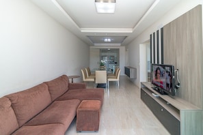 Een moderne woonkamer met een hoekbank, een eethoek met een glazen tafel, en een tv die aan een houten paneel is bevestigd.