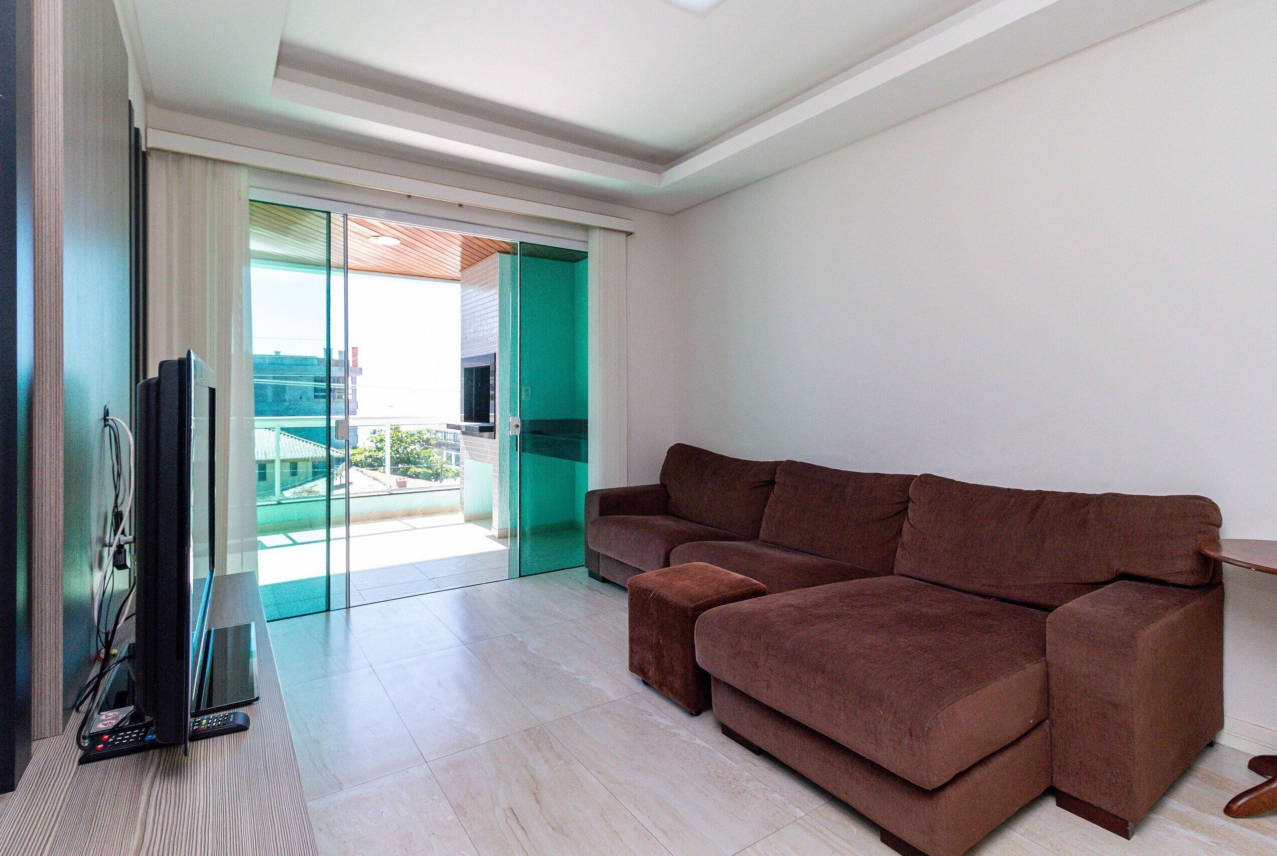 A living room with a brown sofa, a flat-screen TV, and a sliding glass door leading to a balcony.