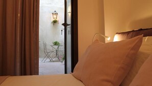 Habitación de hotel con cama, ventana con cortinas y vistas a un área exterior con mesa y sillas, además de una planta colgante.