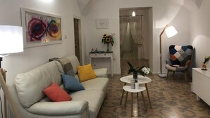 Una sala de estar con un sofá blanco, cojines de colores, una mesita redonda con un jarrón y un cuadro en la pared.