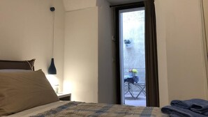 Un dormitorio con cama, mesita de noche, lámpara y vistas a un balcón exterior con una silla y una planta en maceta.