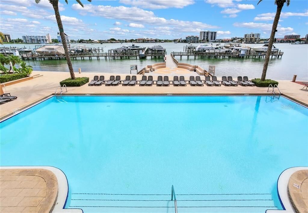 A swimming pool with lounge chairs and a view of a marina with boats and buildings in the background.